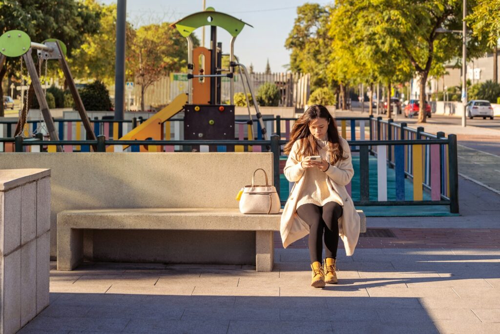 An asian woman sitting on a street bench using her smartphone. Park kindergarten